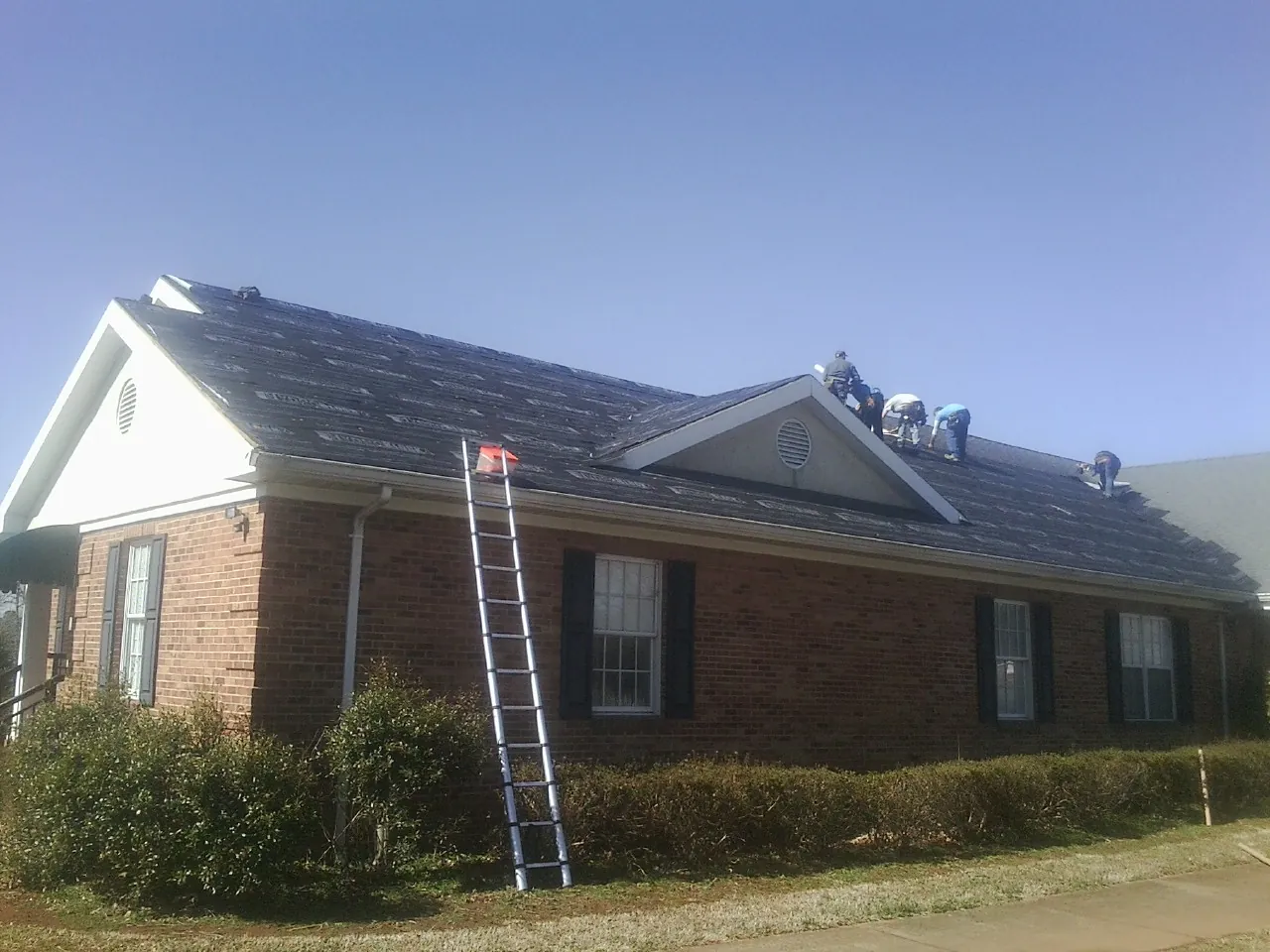 Crew tearing shingles off a brick ranch roof with a ladder set — Emergency Roof Repair work by our crew in Pingree