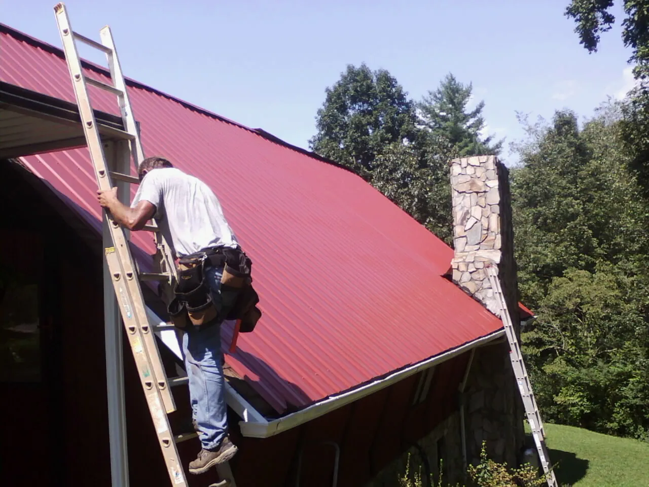 Roofer on a ladder beside a red metal roof and stone chimney — Soffit & Fascia Repair by Roofing Brothers in Pingree